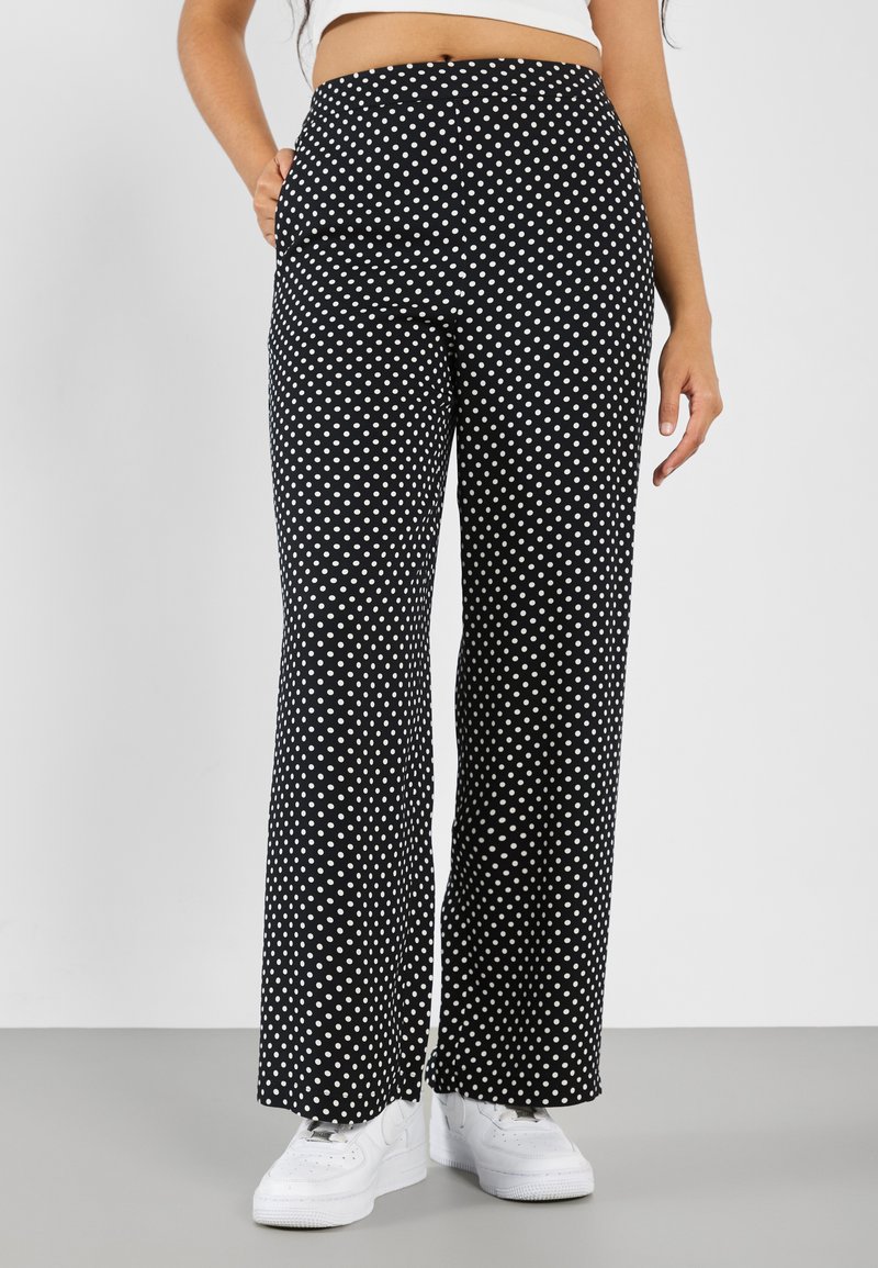 Black wide-leg pants with white polka dots, featuring a high waistband and side pockets, paired with white sneakers on a gray floor.
