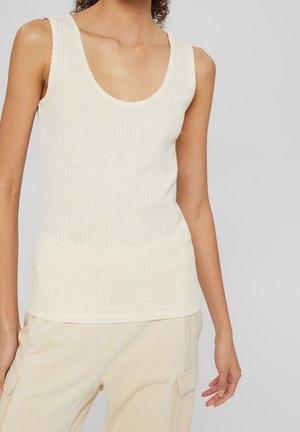 Femme portant un haut sans manches en maille côtelée crème et un pantalon cargo beige clair, debout devant un fond uni gris clair.