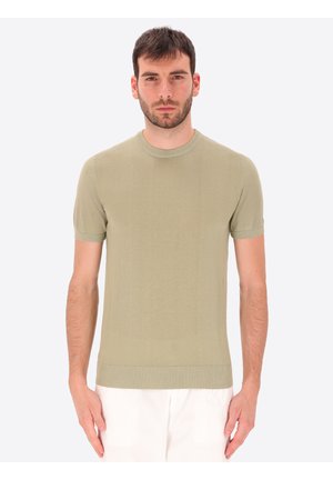 Uomo con capelli corti e scuri che indossa una maglia a maniche corte di colore oliva chiaro e pantaloni bianchi, in piedi davanti a uno sfondo chiaro e uniforme.