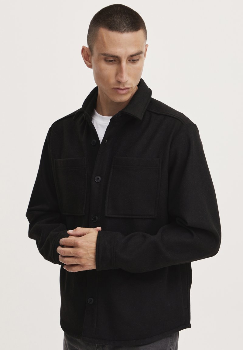Chaqueta negra de manga larga y botones, hecha de tela texturizada. Cuenta con dos bolsillos en el pecho y un collar clásico. Diseño simple y corte recto.