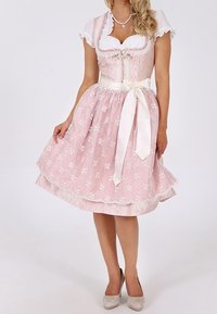 Femme portant une robe traditionnelle dirndl rose clair avec une broderie florale blanche, un chemisier blanc et un ruban de taille blanc noué en nœud.