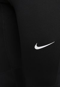 Leggings de sport noirs avec une texture lisse, présentant un logo Nike blanc bien visible sur la cuisse et des détails de couture contrastants.