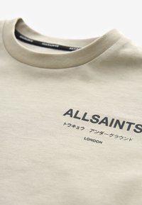 Bézs kereknyakú póló fekete „ALLSAINTS” felirattal, japán karakterekkel és a „LONDON” felirattal a mellrészen.