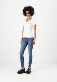 White v-neck t-shirt with a small black logo, blue skinny jeans, and black platform clogs. Smooth texture, classic fit, and minimalist design.