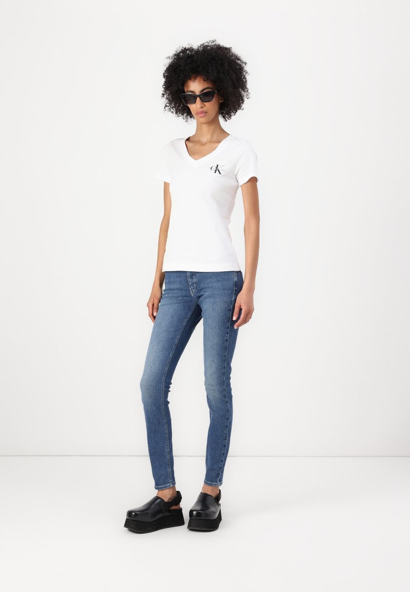 White v-neck t-shirt with a small black logo, blue skinny jeans, and black platform clogs. Smooth texture, classic fit, and minimalist design.