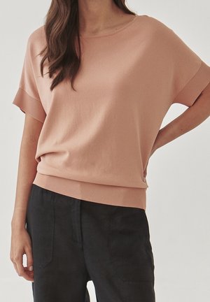 Vrouw die een losse, lichtbruine top met korte mouwen en geribbelde randen draagt, samen met een zwarte broek, staand met één hand op haar heup.