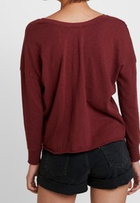 Burgundowa koszula z długim rękawem o luźnym kroju, szerokim dekolcie i podwiniętych mankietach, w zestawieniu z czarnymi szortami dżinsowymi z tylnymi kieszeniami.