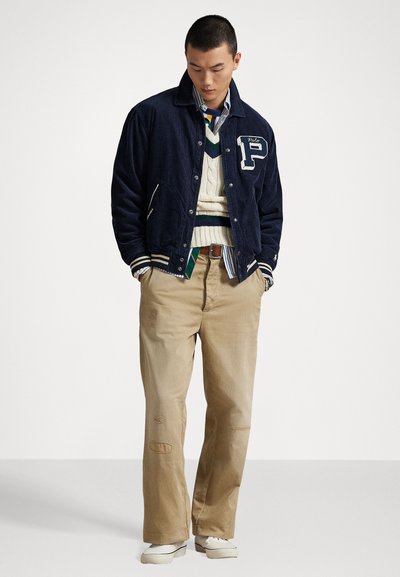 Navy corduroy varsity jacket with white letter detailing, layered over a cream cable-knit sweater and plaid shirt; paired with beige pants and sneakers.