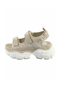 Sandales sport beige avec des straps Velcro, tige texturée, semelle épaisse blanche et support de cheville rembourré, conçues pour le confort et la stabilité.