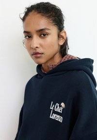 Felpa con cappuccio blu navy realizzata in tessuto morbido; presenta un design a strati con una camicia a quadri sotto e testo ricamato sul davanti.