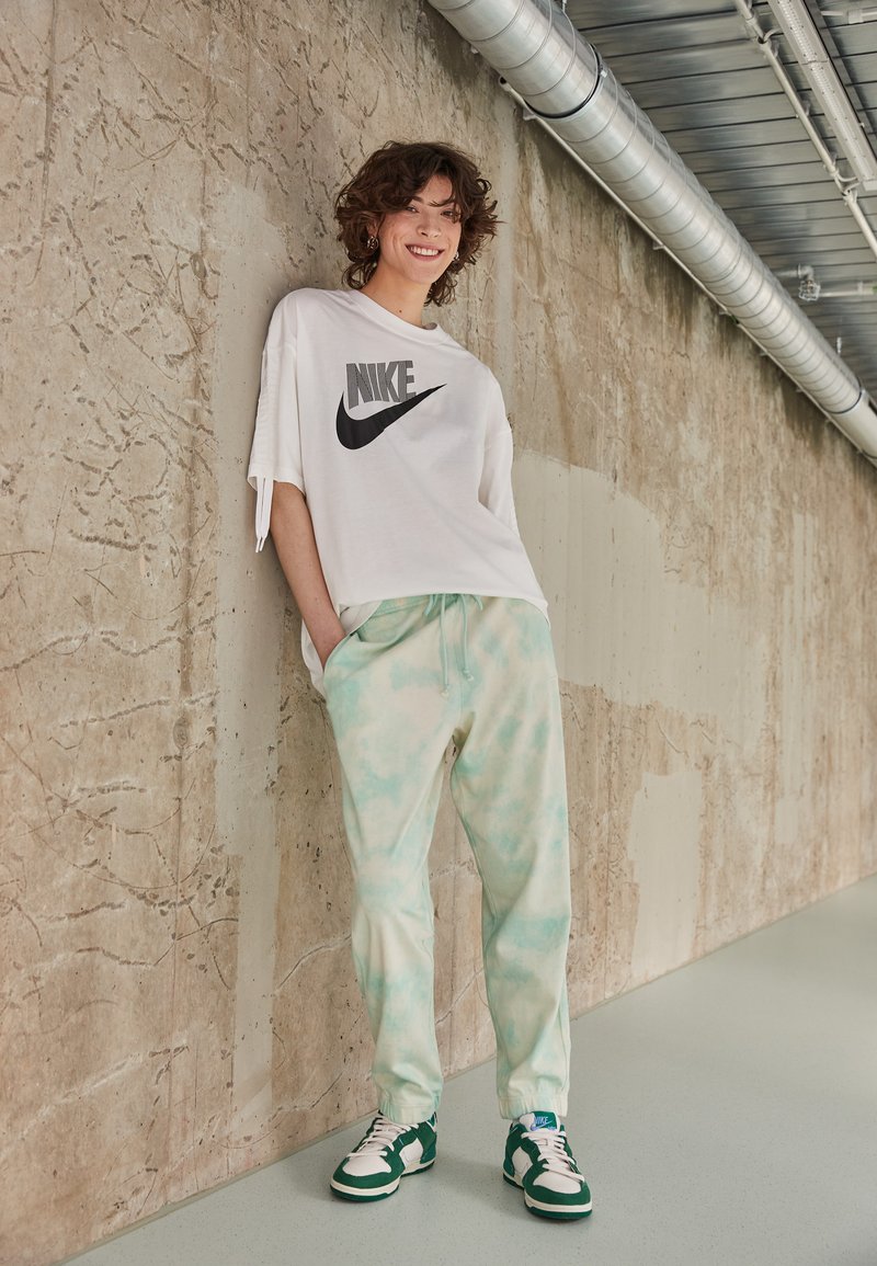 Camiseta blanca de gran tamaño de Nike con logo negro, pantalones de chándal de tie-dye verde menta y zapatillas verdes y blancas, contra una pared de concreto texturizada.