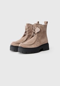 Bocanci de glezna din suede bej, cu șireturi frontale și o talpă texturată din cauciuc negru. Prezintă un accent de marcă pe lateral și o buclă în spate.