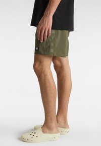 Shorts de bain vert olive avec un motif à carreaux sur le côté gauche, arborant une étiquette de logo noire Vans. Assortis à des chaussures slip-on beige.