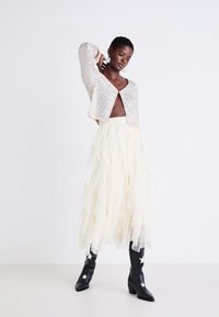 Srebrny kardigan z cekinami, krótki, noszony na warstwowej spódnicy z tiulu w kolorze kremowym. Modelka ma na sobie czarne botki na obcasie z białymi akcentami w kształcie gwiazdy.