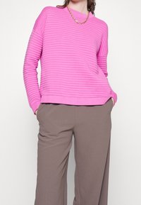 Rosa ribbad tröja med långa ärmar och rund halsringning, kombinerad med bruna vida byxor. Guldhalsbandet lägger till detaljer.