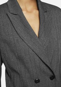 Grijze houndstooth blazer met een geknipt kraag, dubbele knopen en twee zwarte knopen. De stof heeft een gestructureerde uitstraling.