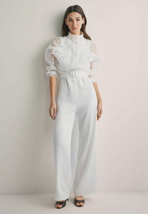 Kobieta ubrana w białą koronkową bluzkę z bufkami na rękawach oraz białe, szerokie spodnie z wysokim stanem i paskiem, stojąca na jasnym, jednolitym tle.