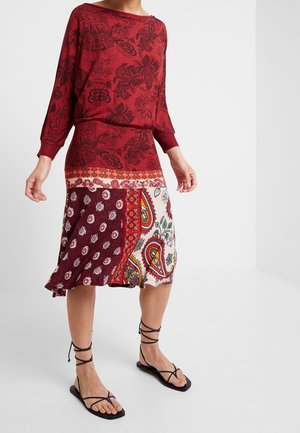 Femme portant un haut à manches longues rouge à fleurs et une jupe patchwork longueur genou avec des motifs cachemire et floraux, assortie à des sandales noires à lanières.