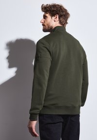 Sweatshirt vert olive avec un col haut et des poignets côtelés, accompagnée d'un écusson sur la manche gauche. Tissu doux et silhouette ajustée.