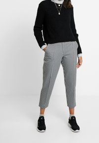 Schwarzer Strickpullover, grauer Rollkragenpullover darunter, kombiniert mit schwarzen und weißen Hahnentrittmuster-Cropped-Hosen und schwarzen Sportschuhen.