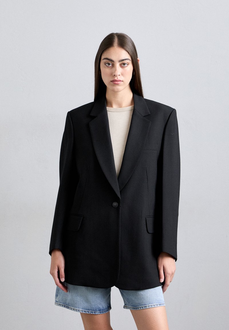 Black blazer with a single button, notched lapels, and two front pockets. Styled over a beige top and paired with denim shorts.