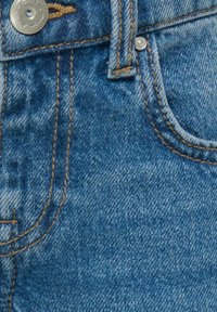 Acercamiento de unos vaqueros de mezclilla azul que muestran el bolsillo delantero, el lazo del cinturón y el botón de metal con detalles de costura naranja.
