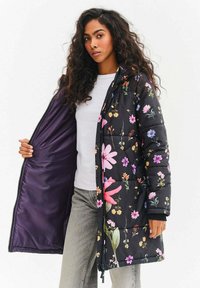 Černý květovaný prošívaný kabát s barevnými květinovými vzory. Má zipové zapínání a fialovou vnitřní podšívku, nošený přes bílou košili.