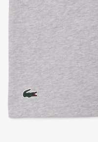 Camiseta de algodón gris con un pequeño logo de cocodrilo verde y acentos rojos en la esquina inferior izquierda. Tela suave y texturizada.