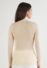 Beżowa bluzka z długim rękawem i kołnierzem typu golf wykonana z miękkiego, lekkiego materiału o dopasowanym kroju i bezszwowym designie na plecach.