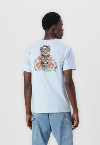 T-shirt de algodão azul claro com uma impressão nas costas de uma máscara tiki rodeada por flores e folhas tropicais coloridas. Corte descontraído, mangas curtas.