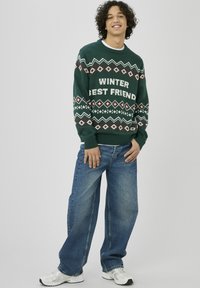 Pull vert en tricot avec le texte "WINTER BEST FRIEND", motifs en losange et étoiles en rouge et blanc. Associé à un jean bleu à jambes larges.