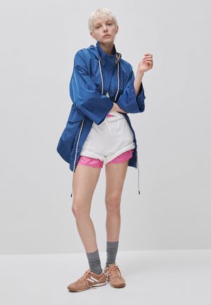 Person trägt eine blaue übergroße Jacke, weiße und pinke Shorts, graue Socken und beige Sneakers und steht vor einem einfarbigen hellen Hintergrund.