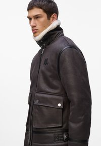 Chaqueta de imitación de cuero marrón oscuro con un alto cuello, borde de borrego, cierre de cremallera y dos bolsillos frontales. Presenta detalles cosidos y un logotipo.