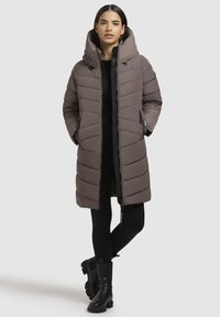 Manteau d'hiver gris long en duvet avec une grande capuche, un design matelassé, des poches zippées et des accents noirs. Porté sur une tenue noire et associé à des bottes noires.