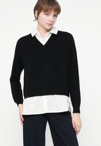 Jeune personne avec des cheveux bruns courts portant un pull noir en V sur une chemise blanche à boutons et un pantalon sombre devant un fond blanc.