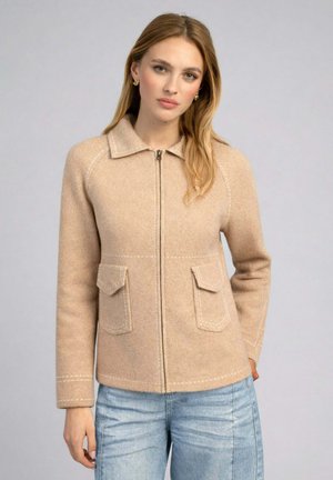 Femme aux cheveux châtain clair porte une veste en laine beige zippée avec col et poches à rabat à l'avant, associée à un jean bleu clair sur fond uni.