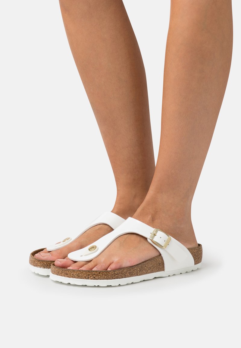 Witte teenslippers met een kurken voetbed, gladde banden en een gouden gesp, voorzien van een gestructureerde zool voor grip.
