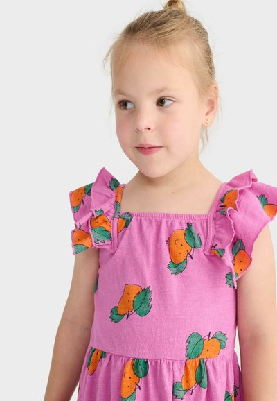 Jeune fille aux cheveux blonds attachés en chignon, portant une robe rose à motifs de fruits orange avec des manches à volants, regardant légèrement sur le côté.
