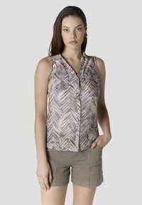 Joven mujer de cabello largo y oscuro, vestida con una blusa sin mangas con botones y estampado, y pantalones cortos beige, de pie frente a un fondo liso.