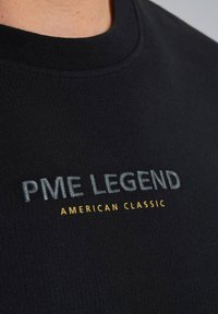 Musta ribattu t-paita, jossa on kirjottu teksti "PME LEGEND" harmaalla ja "AMERICAN CLASSIC" keltaisella, pyöreällä kaula-aukolla.