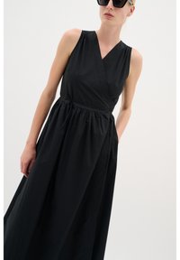 Schwarzes ärmelloses Kleid mit Wickeldesign, gerafftem Taillenbund und Seitentaschen. Hergestellt aus leichtem Stoff mit glatter, matter Oberfläche.