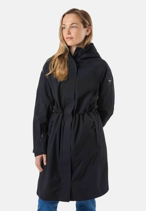 Donna con capelli chiari che indossa un lungo cappotto nero con cappuccio e vita aderente, e jeans blu, in piedi davanti a uno sfondo semplice.