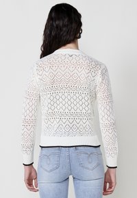 Cárdigan blanco de punto con un patrón de encaje abierto, dobladillo y puños negros acanalados. Diseñado con un motivo de diamante, que ofrece un aspecto texturizado.