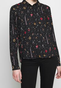 Camicia nera a maniche lunghe con gioielli multicolori e stampe di stelle, indossata con pantaloni neri, mostrando parzialmente il corpo superiore e le braccia.