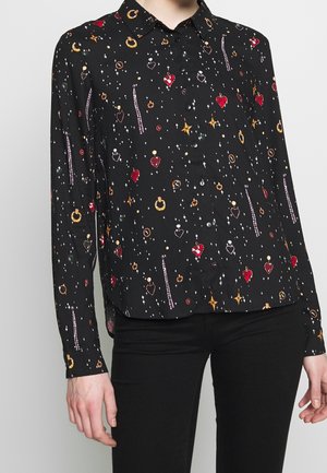 Schwarze Button-Down-Bluse mit mehrfarbigem Schmuck und Sternenmustern, getragen mit schwarzen Hosen, wobei der Oberkörper und die Arme teilweise zu sehen sind.