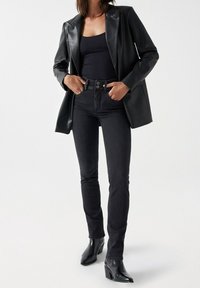 Kvinna klädd i figursydda svarta jeans, svart linne, svart läderblazer och svarta klackboots, stående med händerna i fickorna.