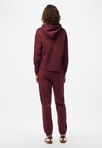 Άτομο με σγουρά μαλλιά που είναι στραμμένο μακριά, φορώντας μπορντό φούτερ, ταιριαστά παντελόνια φόρμας και λευκά αθλητικά παπούτσια με μπορντό λεπτομέρειες σε λευκό φόντο.