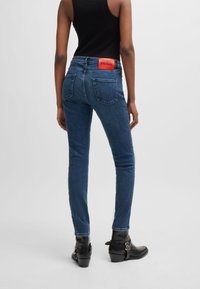 Jeans ajustados de denim azul oscuro con un parche rojo del logo de HUGO en la parte trasera. Cuenta con dos bolsillos traseros y un corte Slim Fit, terminando por encima de unas botas negras hasta el tobillo.