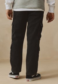 Black cargo pants with side pockets, straight leg design, slight texture, paired with black and white sneakers. Background is neutral.