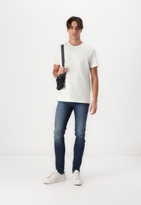 Bílá bavlněná tričko s embosovaným logem, modré slim-fit džíny a bílé tenisky. Černá crossbody taška se strapcem. Jednoduchý a moderní design.